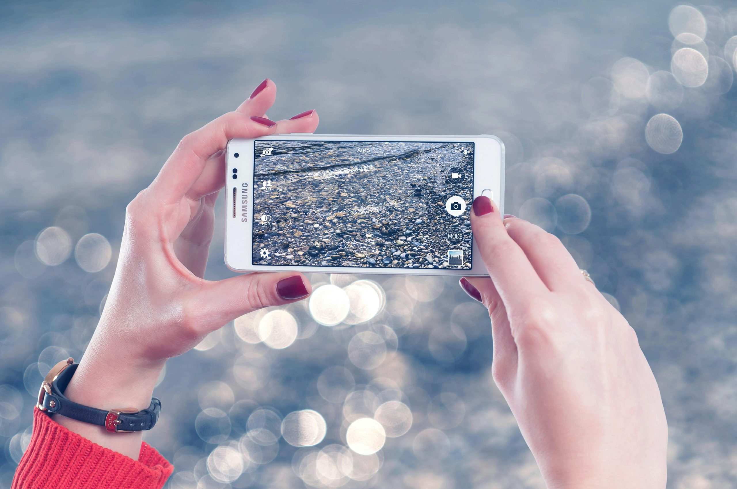 Mains tenant un smartphone pour prendre une photo au bord de l’eau avec un arrière-plan flou et lumineux