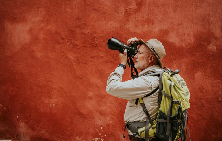 Photographe en voyage avec sac à dos et téléobjectif devant un mur rouge, pour illustrer le choix d’un objectif photo voyage