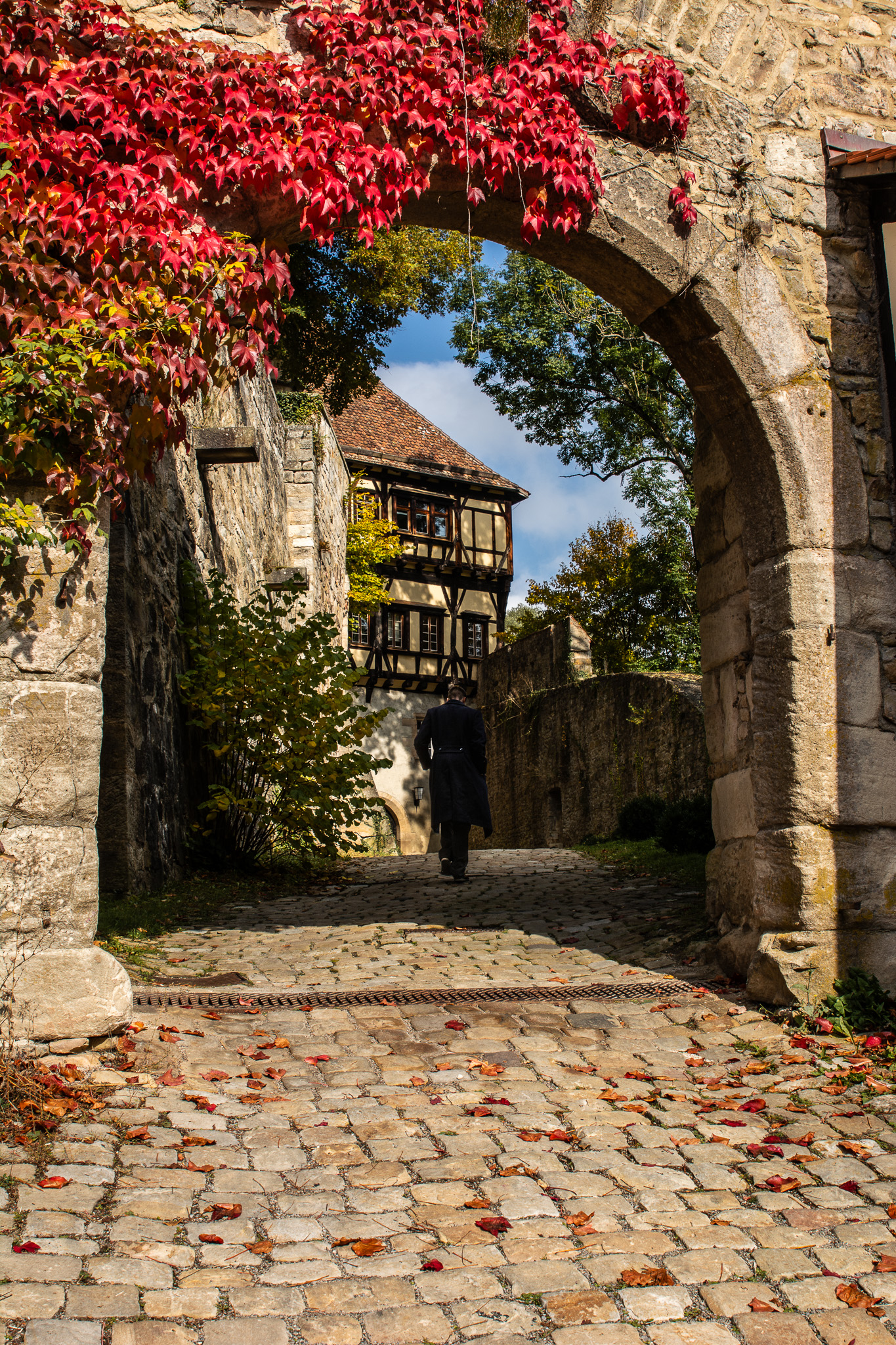 Une personne traverse une arche en pierre bordée de feuilles rouges, devant une maison à colombages.