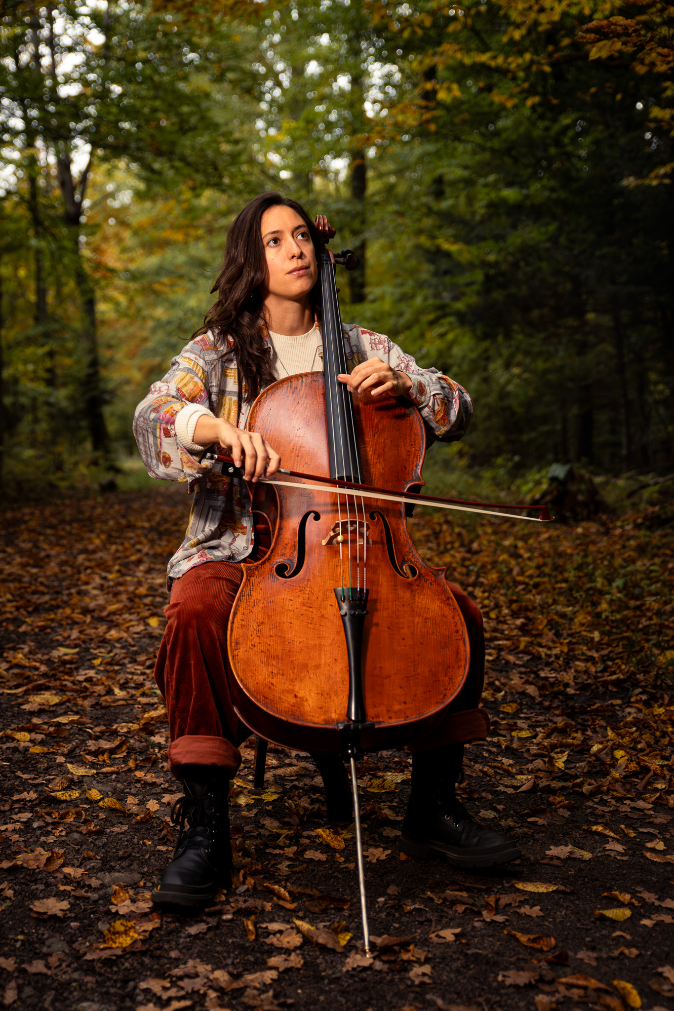 La violoncelliste Hannah joue dans une forêt au feuillage vert et doré, sur un sol couvert de feuilles mortes.